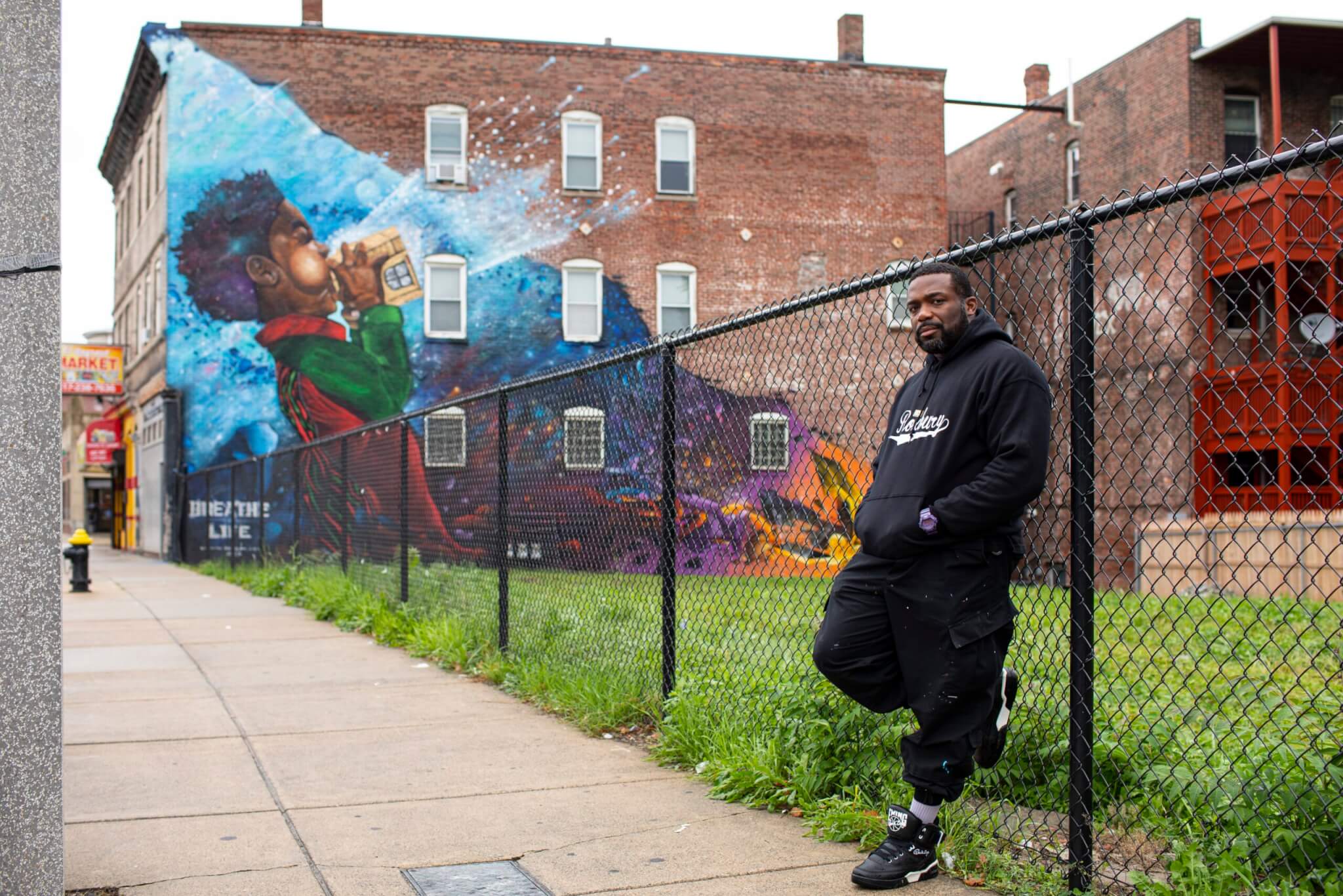 An artist stands by a fence in front of a colorful mural, in the neighborhood of Dorchester.