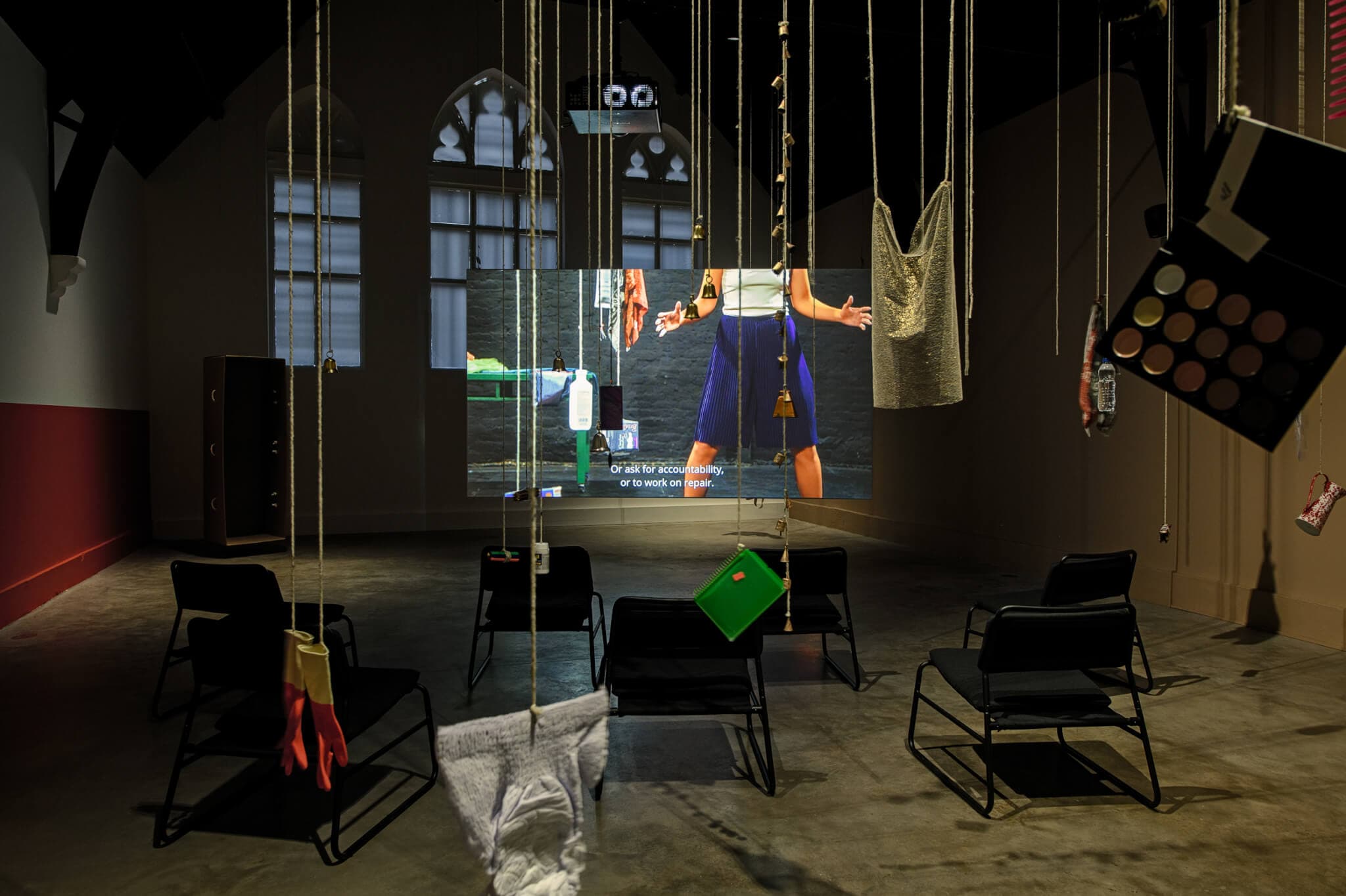 In a large room, everyday objects—such as rubber gloves, eyeshadow palettes, notebooks, bells, and water bottles—dangle from strings suspended from the ceiling. In the background, a video piece is projected onto a screen, with chairs scattered in front of it.
